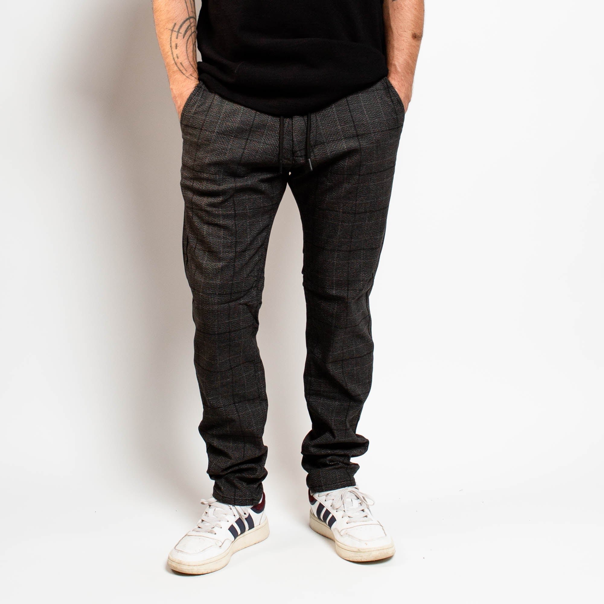 Person wearing dark plaid pants and white sneakers against a plain background By Projek Raw