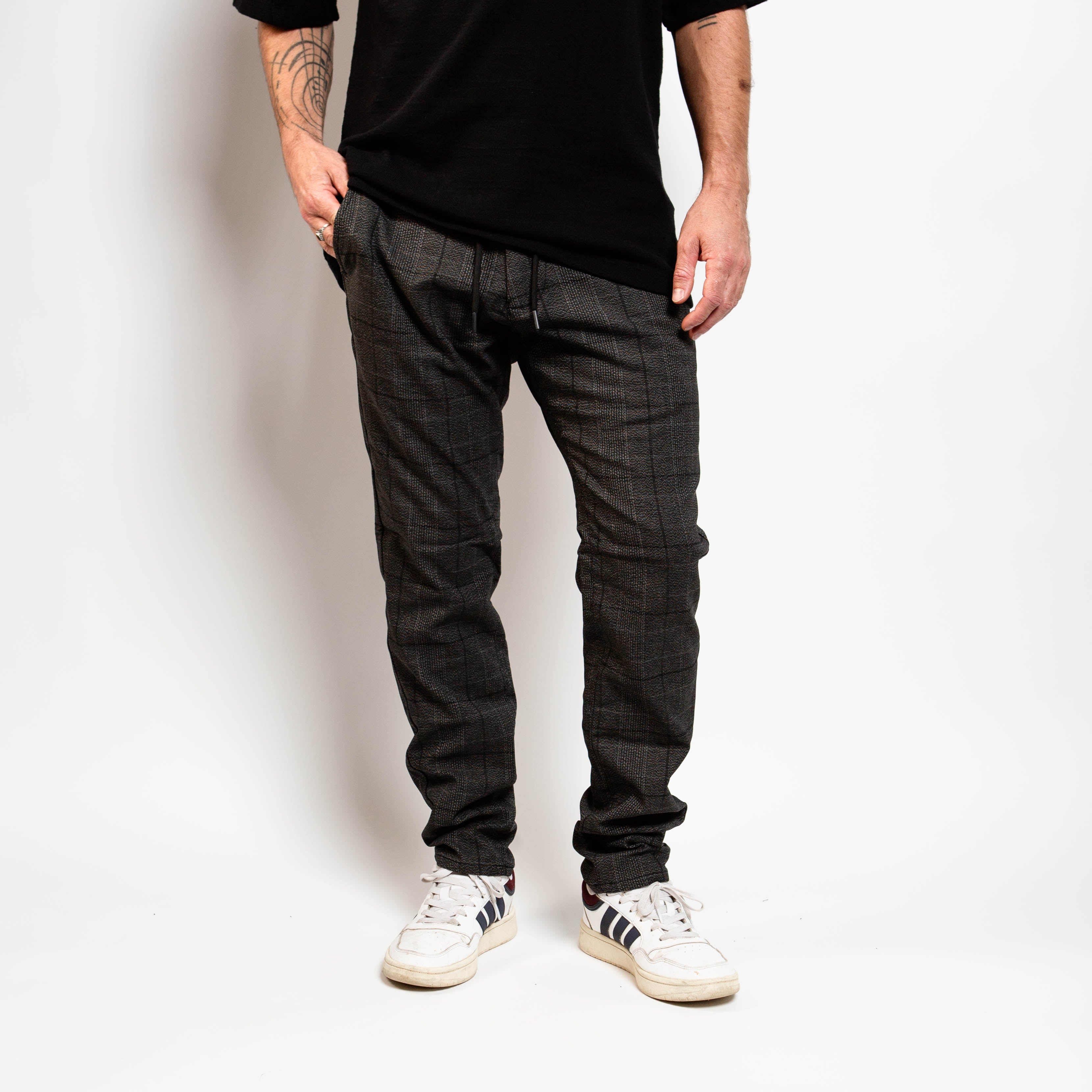Person wearing dark plaid pants and white sneakers against a plain background By Projek Raw