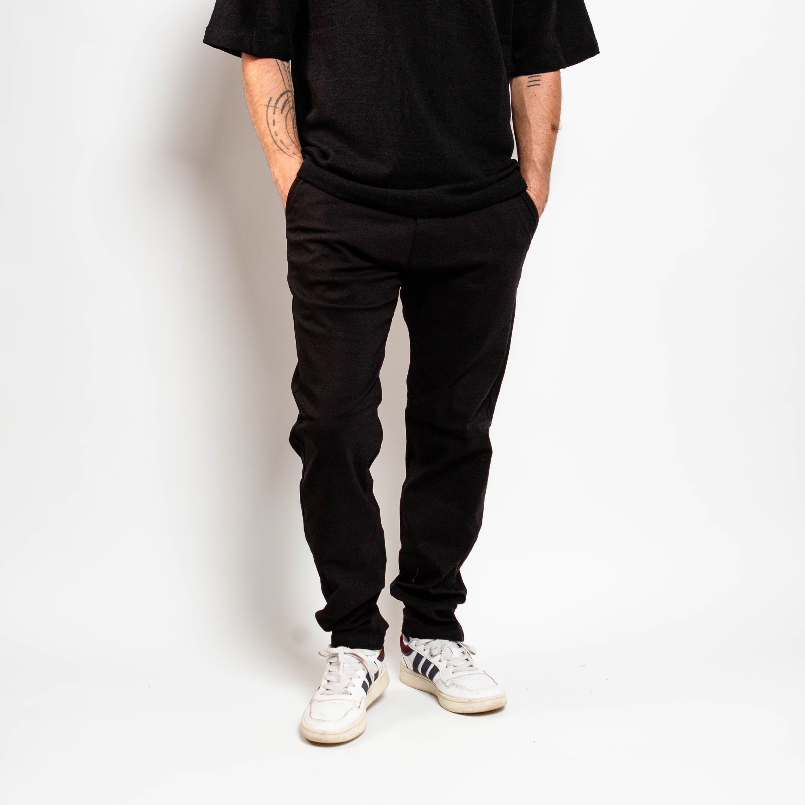 Person wearing a black t-shirt, black pants, and white sneakers against a white background By Projek Raw