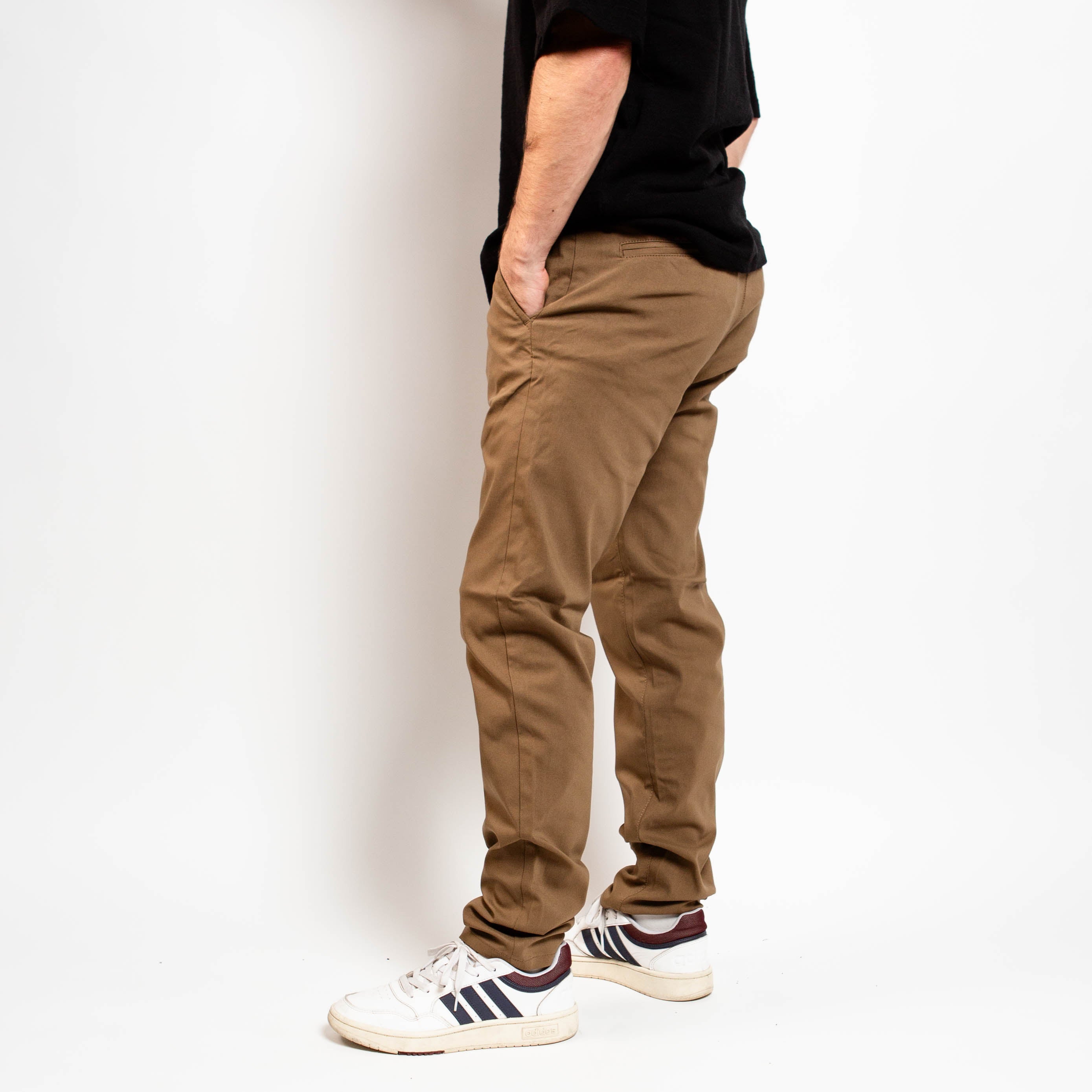 Person wearing brown pants and white sneakers with black stripes on a plain background By Projek Raw