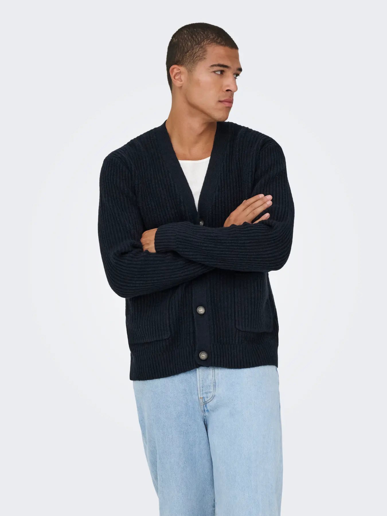 Man wearing a dark blue cardigan over a white shirt with light blue jeans on a plain background by only & sons