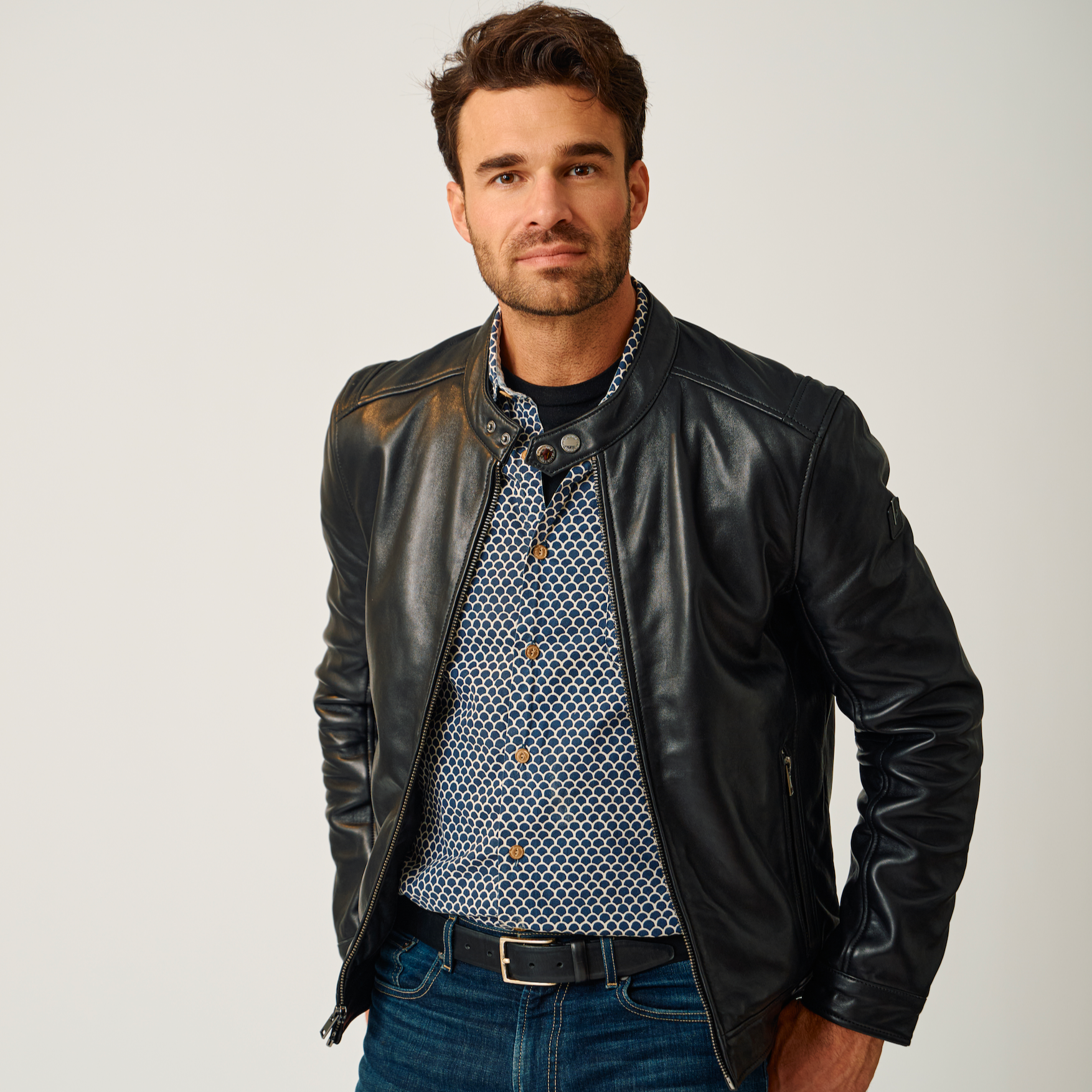 Man wearing a black leather jacket, patterned shirt, and blue jeans on a white background