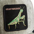 Patch of a praying mantis with 'HEARTBREAKER' text on a black background, attached to a gray fabric surface.