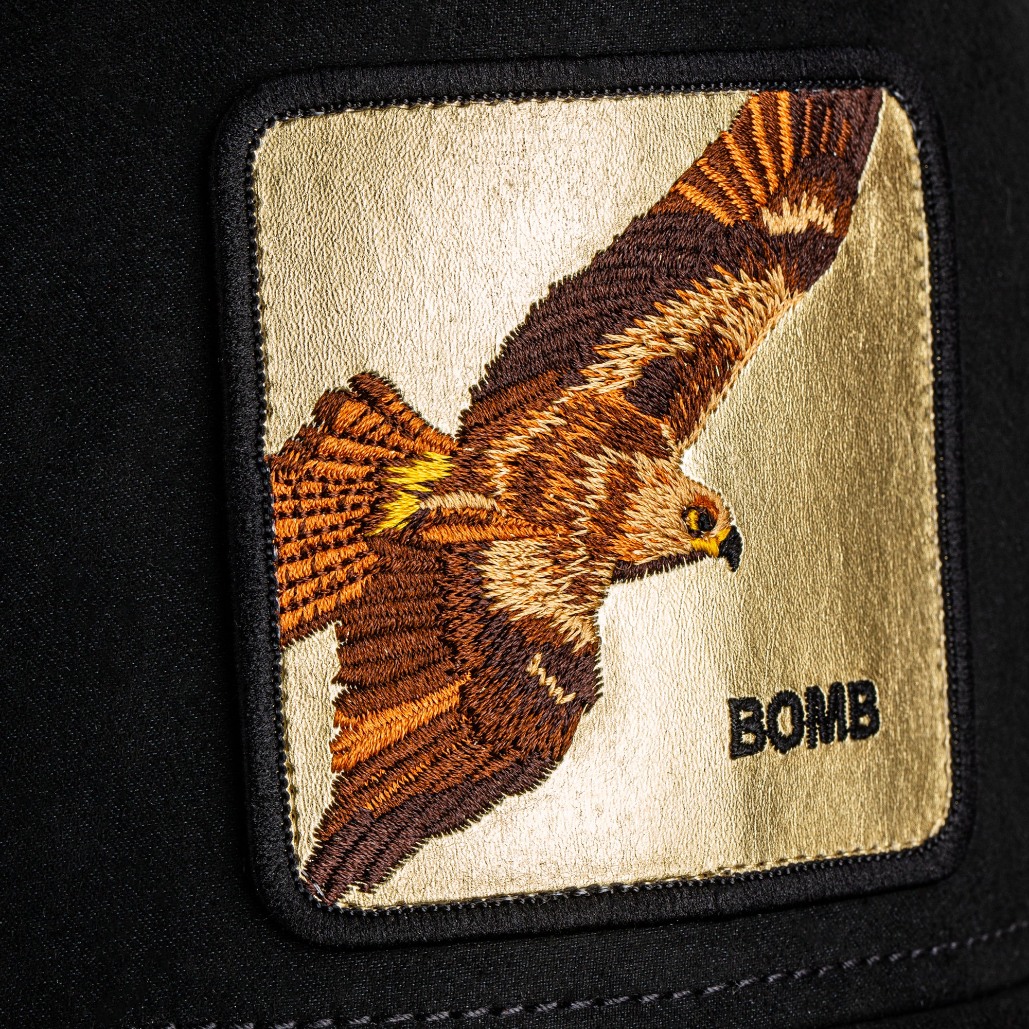 Embroidered patch of a hawk with 'BOMB' text on a beige background, attached to a black surface.