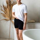 A man wearing a white v-neck t-shirt and black boxer briefs stands barefoot in a bright, modern bathroom next to a white freestanding tub.