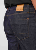 Close-up of dark blue jeans with a visible brand label on a light gray background By Duer Jeans