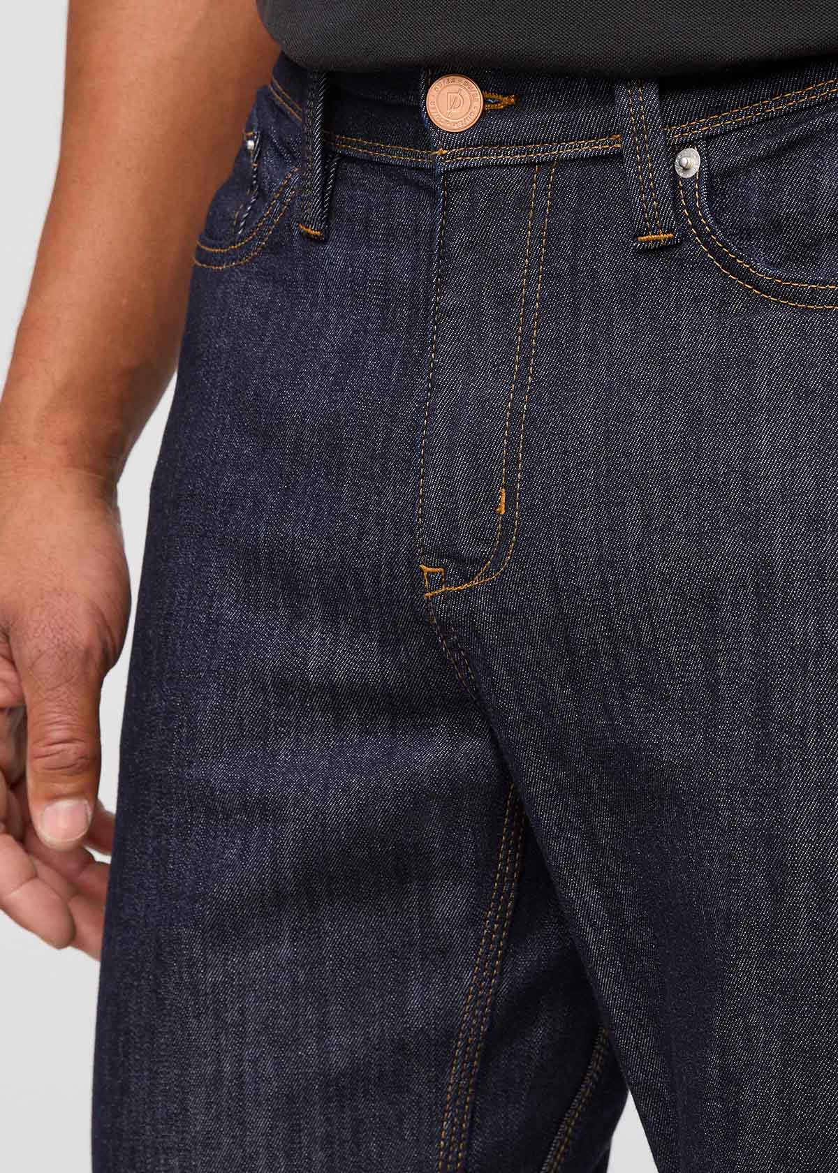 Close-up of dark blue jeans with a visible button on a light gray background By Duer Jeans