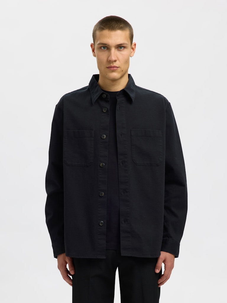 Male model wearing a black Twill overshirt by SELECTED Homme, featuring button-up front, two chest pockets, and a relaxed minimalist fit.
