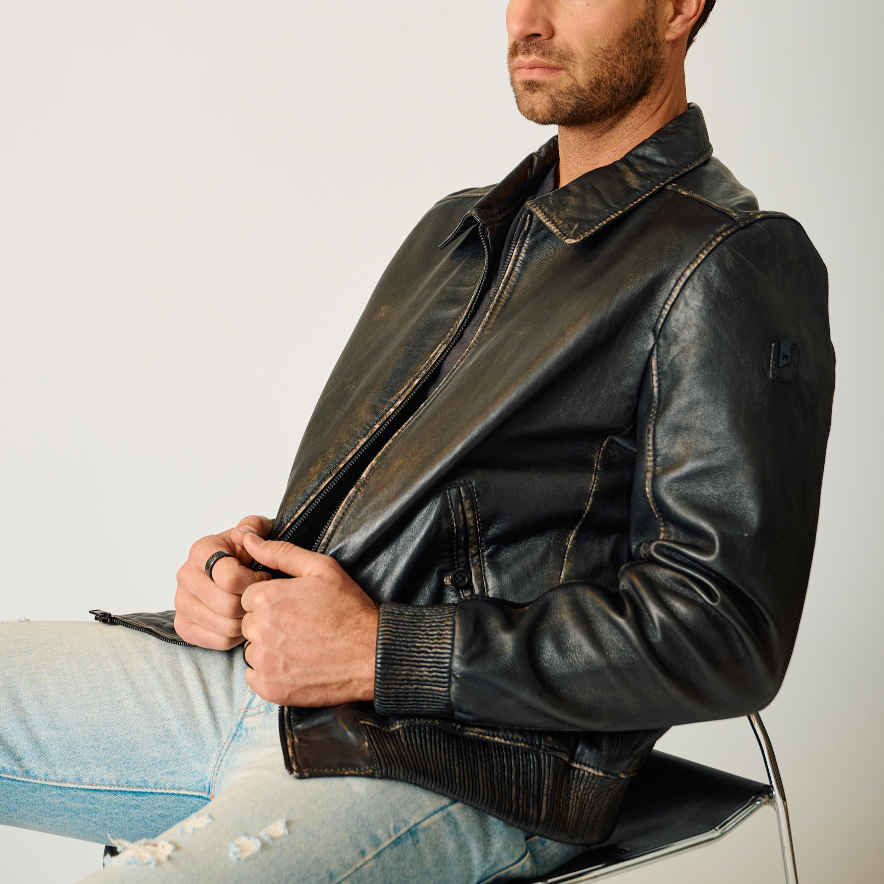 Man wearing a black leather jacket sitting on a white chair against a light gray background